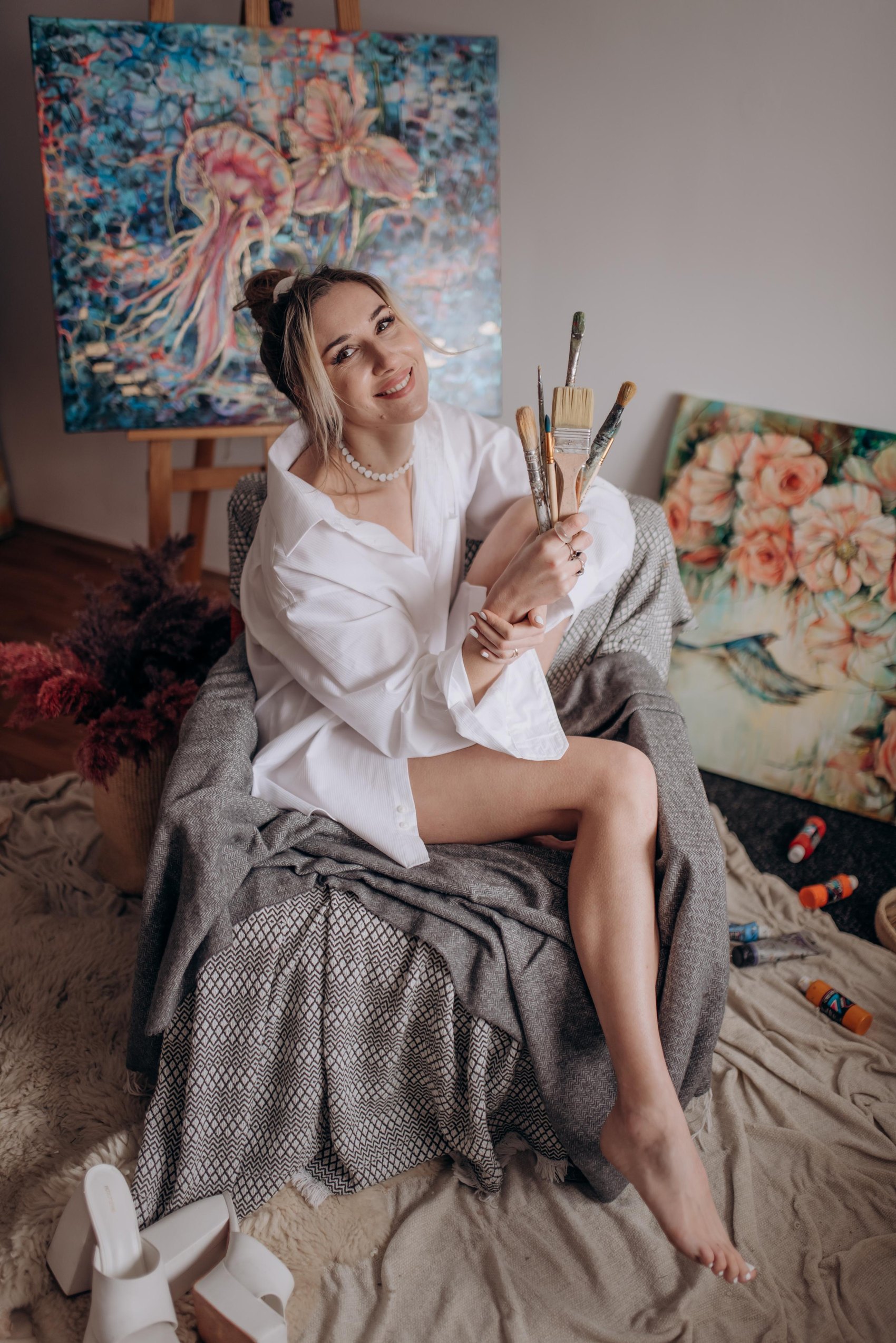 Liudmyla holding paintbrushes in her studio, surrounded by her paintings